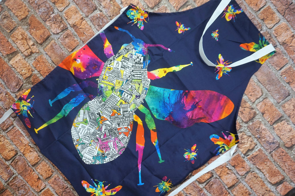 Picture of a navy blue cotton apron printed with a large colourful bee containing an abstract manchester skyline design
